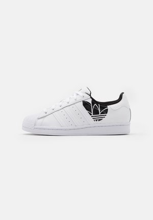 Biała tenisówka Adidas z trzema bocznymi paskami, czarną wyściółką i czarnym logo trefoil na części piętowej, sfotografowana na białym tle.