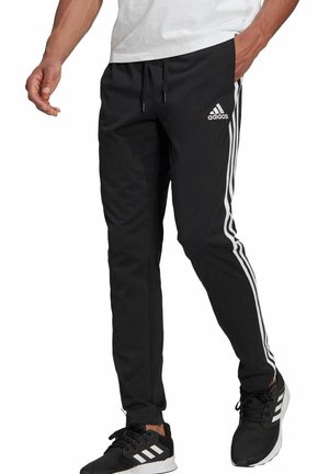 Man draagt zwarte Adidas trainingsbroek met witte strepen aan de zijkanten en zwarte Adidas sneakers met witte zolen.