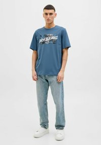 Blaues Baumwoll-T-Shirt mit Grafikdruck, kombiniert mit hellblauen Jeans und weißen Sneakers. Kurze Ärmel und lockerer Schnitt.