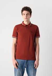 Polo tričko v hrdzavej farbe s rebrovaným golierom s bielymi a červenými pruhmi, krátkymi rukávmi a čiernym logom na hrudi.