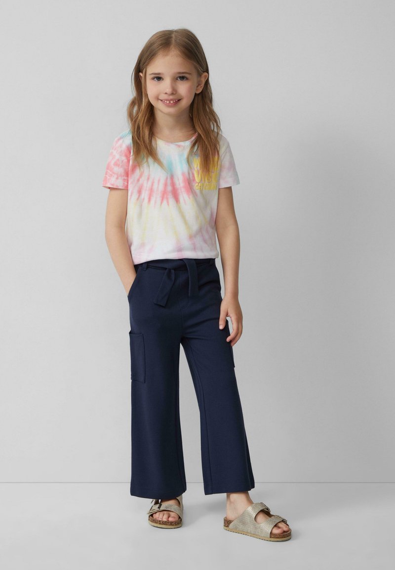 Tie-dye t-shirt met korte mouwen in roze, geel en blauw met de tekst "Sweet Growing", gecombineerd met marineblauwe wijde broek en lichte sandalen.