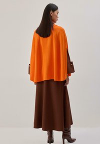 Cape tricotée orange à coupe ample, superposée à une longue jupe marron. Dotée de manches ajustées avec un détail à bouton.