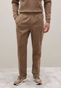 Braune elastische Hosen mit seitlichen Taschen, schmal zulaufendem Design und glatter Textur, kombiniert mit passenden braunen und grauen Sneakers.