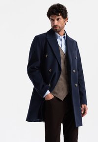 Cappotto di lana blu navy con bottoni doppi, ampie rever, e vestibilità sartoriale, indossato su un gilet e una camicia a motivi houndstooth marroni.