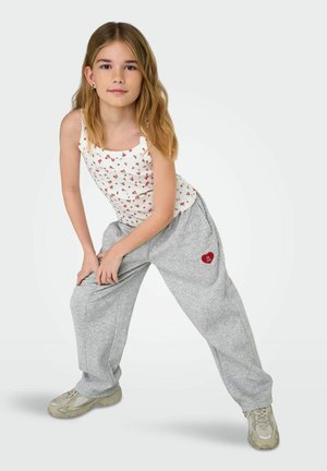 Mladi deklica z dolgimi blond lasmi, oblečena v belo top z rožnatim vzorcem in sive športne hlače z rdečim obližem v obliki srca, pozira s položaji rok na kolenih.