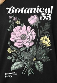 Svart T-shirt med en blommig design med rosa, gula och gröna blommor, texten "Botanical 55" och "Beautiful Story" i vitt.