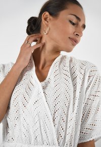 Een vrouw met haar haar in een knot draagt een witte, met kantpatroon versierde top. Ze raakt haar nek voorzichtig aan en laat haar oorbellen en een ring zien.