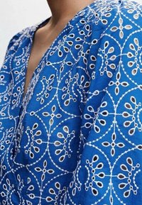 Indumento blu ricamato con intricati motivi floreali bianchi. Il tessuto ha una consistenza morbida, con un design a V e una vestibilità leggermente ampia.