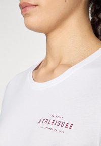 Biely tričko vyrobené z bavlny, s okrúhlym výstrihom a potlačeným červeným logom s nápisom "ATHLEISURE" v blízkosti hrudníka.