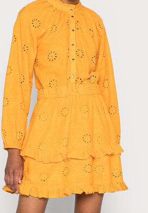 Femme portant une robe jaune vif à manches longues, motifs floraux découpés, accents à volants, et boutons sur le devant.