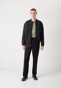 Veste bomber noire avec une surface texturée, t-shirt vert en dessous, pantalon noir et baskets sombres. Design minimaliste et coupe moderne.