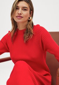 Sarkanais adīts kleita ar ribotu tekstūru, augstu apkakli un garām piedurknēm, kombinēta ar apaļām zelta auskaru. Fons ir brūns krēsls.