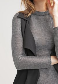 Gilet senza maniche nero sopra un top grigio aderente a maniche lunghe con un motivo orizzontale texturizzato. Il gilet ha un colletto morbido e una chiusura aperta.