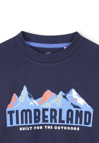 Sötétkék pamut póló, stilizált kék, fehér és narancssárga hegyeket ábrázoló grafikával, alatta "TIMBERLAND" felirattal.