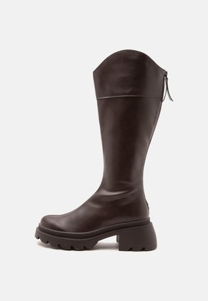 Botte haute en cuir brun foncé avec une semelle épaisse, un talon bloc et une fermeture éclair à l'arrière, photographiée sur un fond neutre.