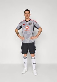 Harmaa jalkapallopaita mustilla yksityiskohdilla, jossa on geometrinen kuvio, punaiset olkapääyksityiskohdat ja logot. Yhdistetty mustiin shortseihin ja valkoisiin sukkiin.