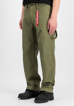 Mann trägt olivgrüne Cargohose mit rotem "Remove Before Flight" Schlüsselanhänger und schwarzen Schnürstiefeln, steht vor einfarbigem Hintergrund.