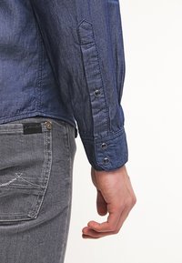 Granatowa koszula dżinsowa z zapięciem na guzik w mankietach, o gładkiej fakturze i subtelnych detalach szycia. W połączeniu z szarymi dżinsami.