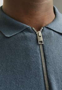 Dunkelblaues, strukturiertes Strickpolo mit kurzem Kragen und silbernem Metallreißverschluss. Nahaufnahme, die den Stoff und die Reißverschlussdetails hervorhebt.