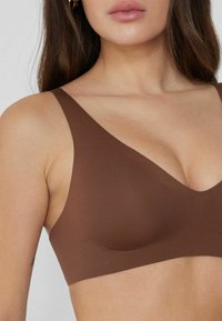 Hnědá bezšvová podprsenka s širokými ramínky a výstřihem do V. Materiál je hladký a matný, což zdůrazňuje minimalistický styl.
