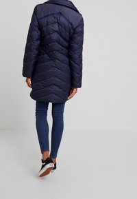 Marinblå puffjacka med zigzag-mönster, lätt material, termisk isolering och krage, tillsammans med blå jeans och svarta sneakers.