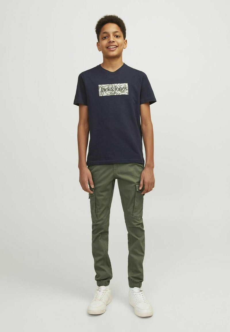 T-shirt bleu marine avec un graphique camouflage et un col rond, associé à un pantalon cargo olive avec des poches latérales et des poignets élastiques, porté avec des baskets blanches.