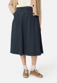 Marineblauwe denim rok met twee voorzakken, een knoopsluiting en een knielange A-lijn ontwerp. Draagt beige sneakers en crèmekleurige sokken.