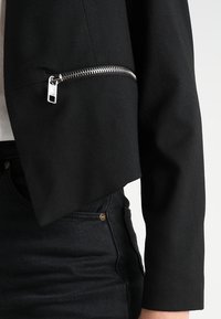 Musta lyhyt takki, jossa hopeanvärinen vetoketju hihassa. Valmistettu sileästä kankaasta, siinä on jämäkkä leikkaus ja minimalistinen muotoilu.