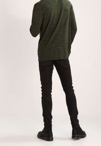 Jersey de punto verde con un acabado texturizado, combinado con jeans negros ajustados y botas negras. La vista es desde atrás, destacando la silueta del atuendo.
