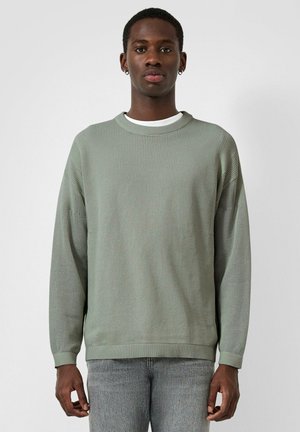 Mężczyzna w jasnooliwkowym swetrze o fakturze, nałożonym na białą koszulę i wyblakłe szare dżinsy, stoi na tle jednolitego białego tła.