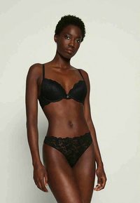 Sieviete ar īsiem cirtainiem matiem, ģērbusies melnā mežģīņu krūšturī un atbilstošās augstās apakšbiksēs, stāv uz viendabīga gaiša fona.