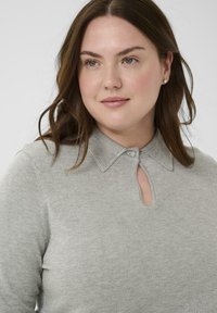 Vrouw met lang bruin haar, gekleed in een grijze trui met een knoopsluiting en een keyhole halslijn, die iets opzij kijkt.