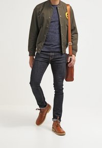 Veste bomber vert olive, pull bleu, jean sombre, bottines marron, sac messager en cuir marron, personne debout devant un fond uni.