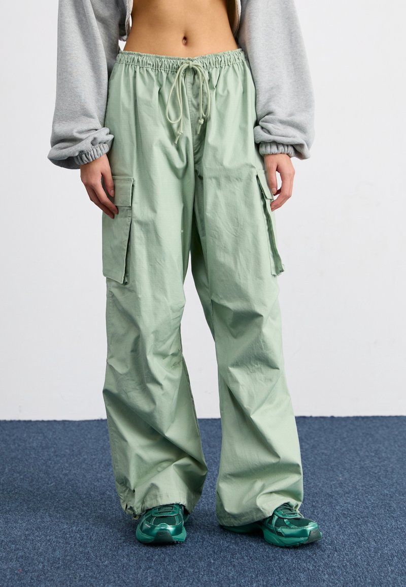 Lysgrønne cargo bukser med snøre i taljen, elastiske manchetter og to sidelommer med klap, parret med grønne sneakers. Glat tekstur.