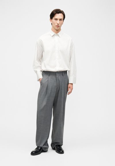 Άνδρας με λευκό ριγέ πουκάμισο με κουμπιά, γκρι παντελόνι φαρδύ με πιέτες και μαύρα παπούτσια φόρμας στέκεται με το ένα χέρι στην τσέπη.
