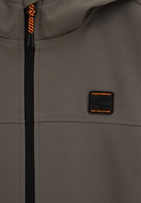 Jachetă gri, tip soft-shell, cu închidere cu fermoar, având șnur portocaliu și o etichetă neagră cu textul „Young Originals” pe piept.