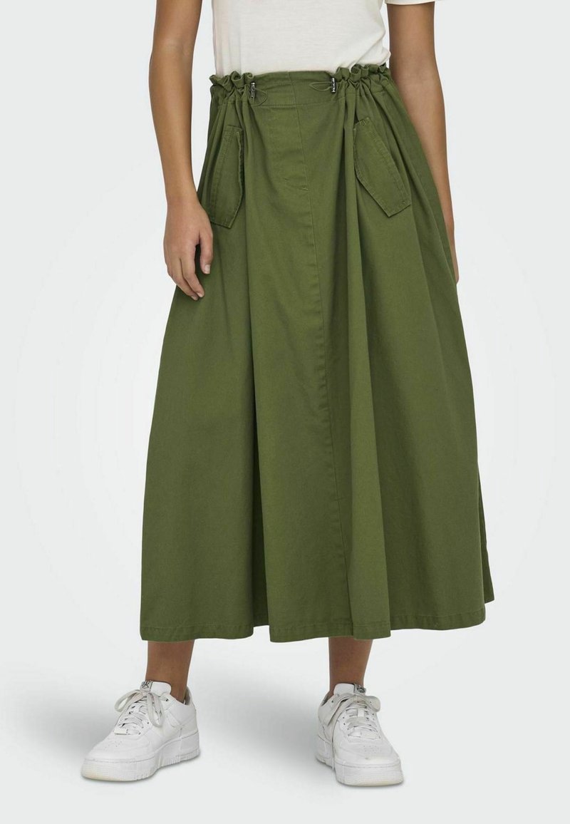 Personne portant une jupe midi olive avec poches avant et taille froncée, associée à des baskets blanches.