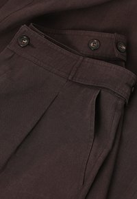Pantalon marron foncé à taille haute, ceinture boutonnée, poches latérales et coutures visibles sur tissu en coton.