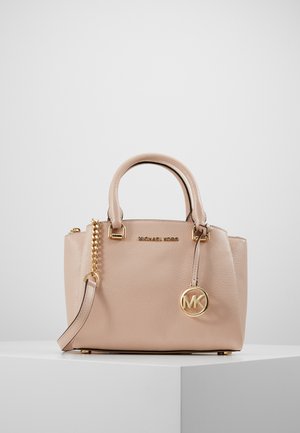 Beige Michael Kors handtas met een korrelige lederen textuur, gouden kettingriem, dubbele handvatten en een ronde MK-logo bedel op een wit voetstuk.