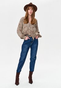 Kobieta w brązowym kapeluszu, bluzce w panterkę, niebieskich dżinsach z beżowym paskiem i brązowych botkach, stojąca z rękami w kieszeniach.