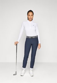Frau in einem weißen Langarm-Polo, mit marineblauen Hose und weißen Sneakers, die einen Golfschläger hält und vor einem schlichten weißen Hintergrund steht.