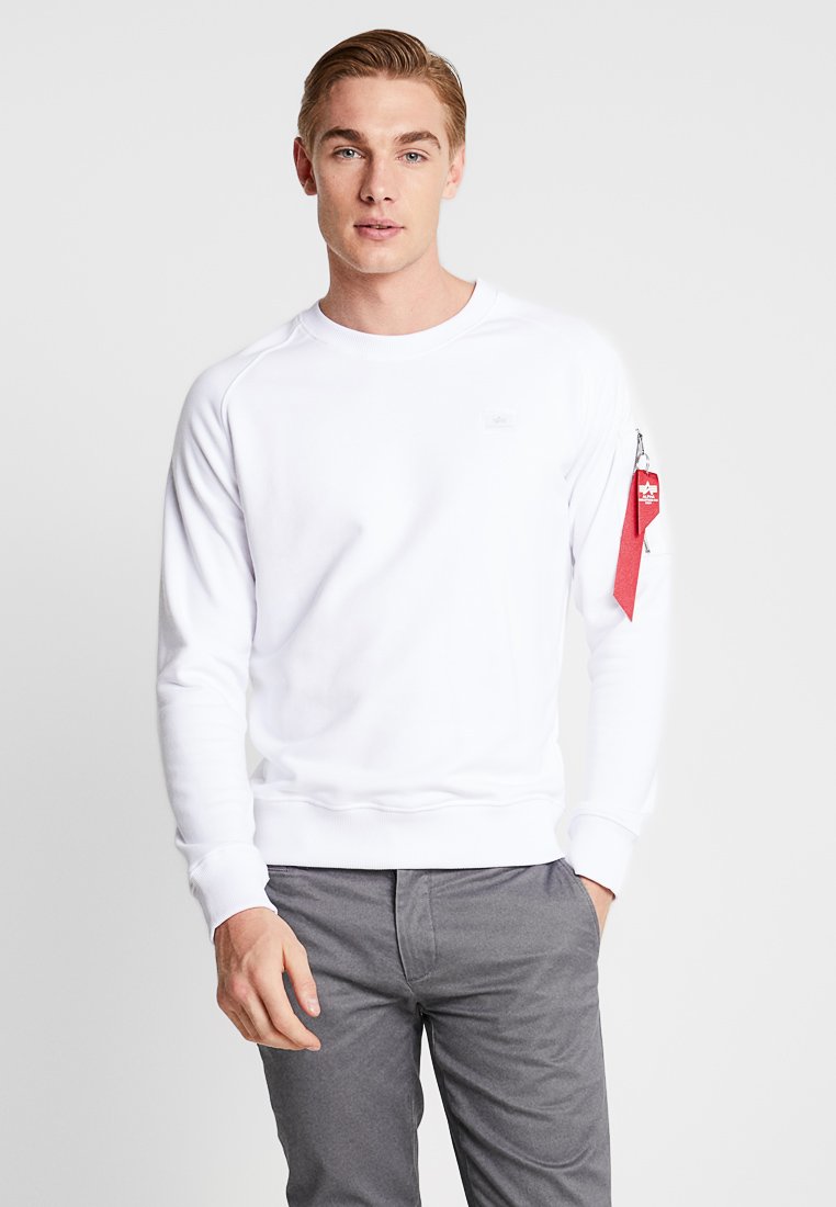 Felpa bianca con collo a giro, polsini e orlo a costine, che presenta un piccolo logo sul petto e un'etichetta rossa sulla manica sinistra. Pantaloni grigi.