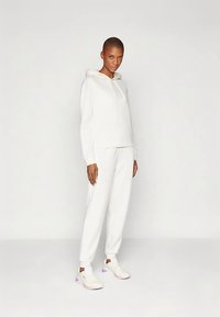 Sweat à capuche blanc avec cordon de serrage, assorti à un pantalon de survêtement blanc fuselé. Matière douce avec une texture lisse. Le modèle porte des baskets blanches.