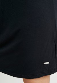 Svart bomullskjol med en slät yta, lätt avsmalnande design och en liten silvrig logotagg nära fållen.