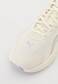 Detailní záběr bílé tenisky Puma v oblasti špičky, která obsahuje síťovinu, tkaničky a modré logo Puma na horní části.