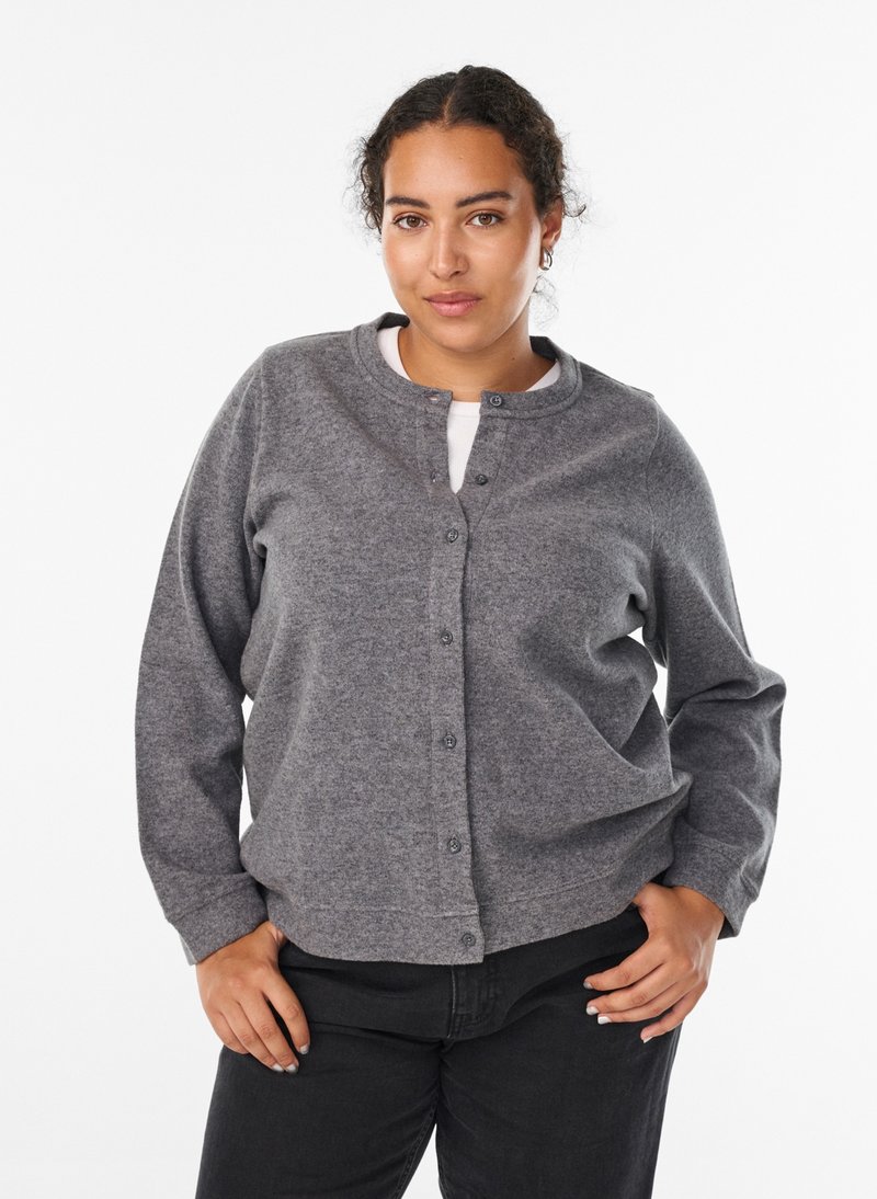Giovane donna con capelli ricci che indossa un cardigan grigio con bottoni, top bianco e pantaloni neri, posa con le mani sui fianchi davanti a uno sfondo bianco.