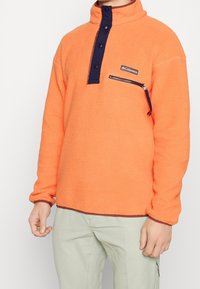 Persona vistiendo un forro polar naranja con botones a presión marinos y un bolsillo en el pecho con cremallera, combinado con pantalones de un beige claro.