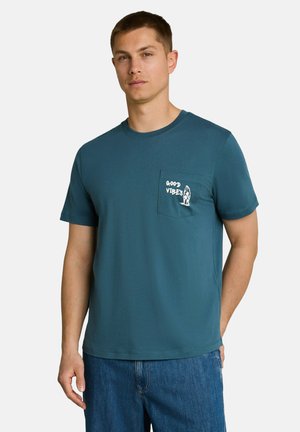 Junger Mann trägt ein türkisfarbenes kurzärmliges T-Shirt mit einer Vorderseite Tasche, die den Text "GOOD VIBES" und eine kleine Surfbrett-Grafik hat.
