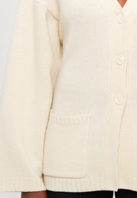 Kremowy sweter z dzianiny z guzikami z przodu, ściągaczami przy mankietach i u dołu oraz małą prostokątną kieszonką po bokach.