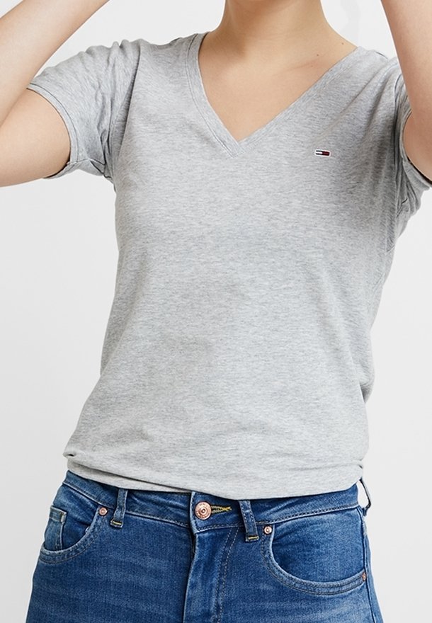 Personne portant un T-shirt gris clair à col en V avec un petit logo sur la poitrine et un jean bleu, torse et taille visibles, bras levés.